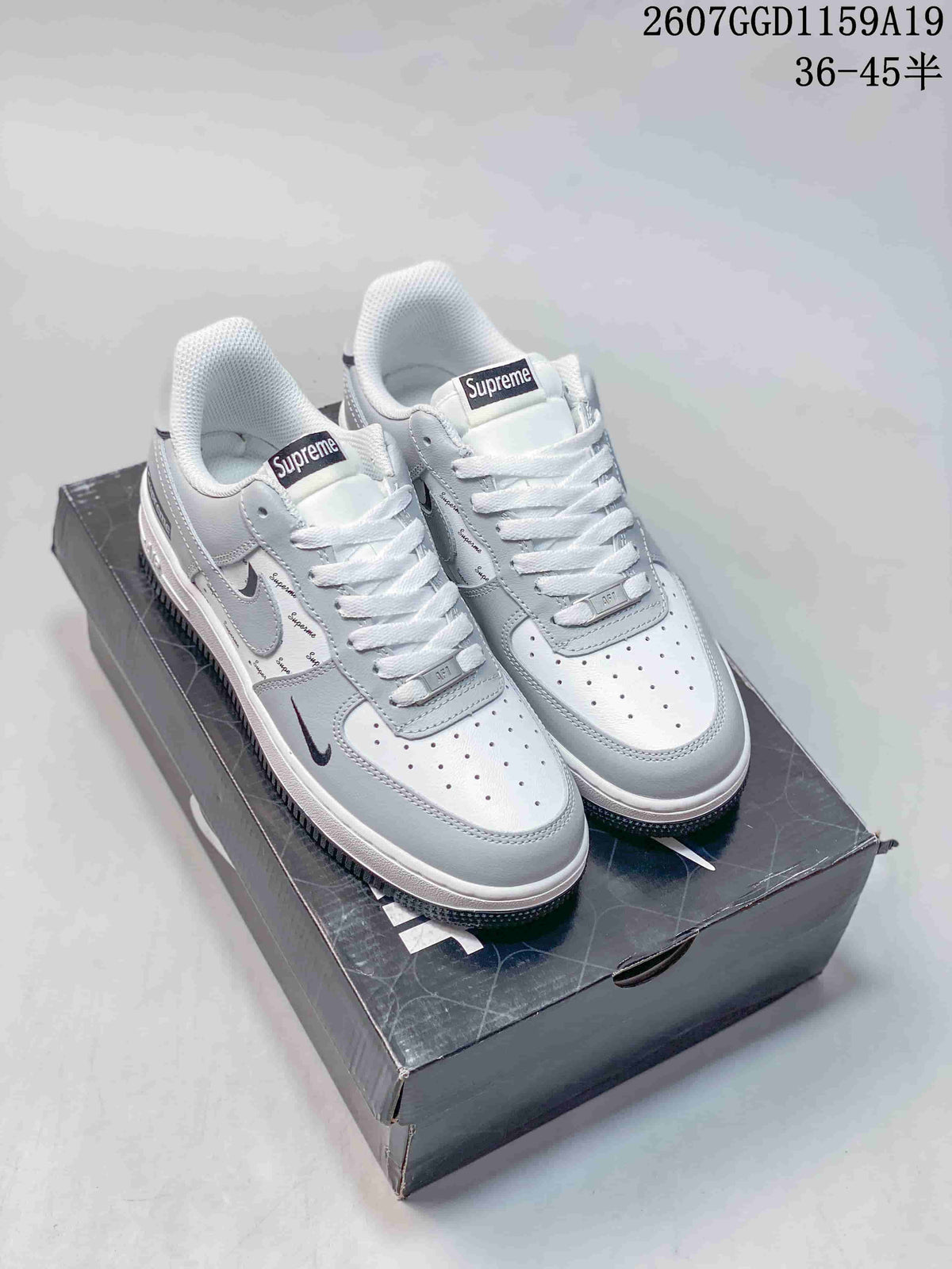 Pair of white low-top sneakers displayed on a shoebox, showing dark Swoosh details and Supreme-branded tongue labels.