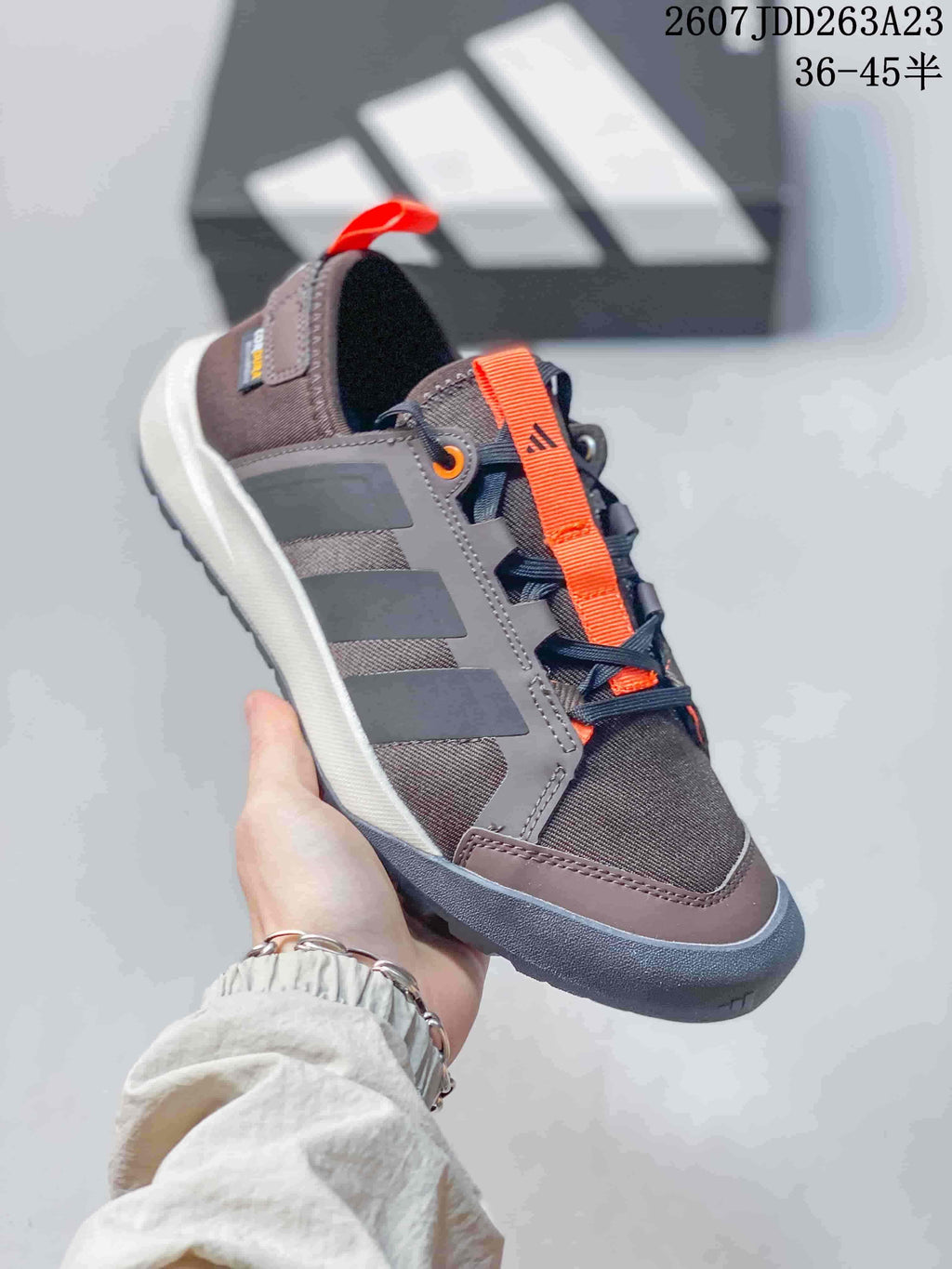 Close side view of a brown Adidas Terrex sneaker with orange lace strap, three-stripe panels and heel pull tab.