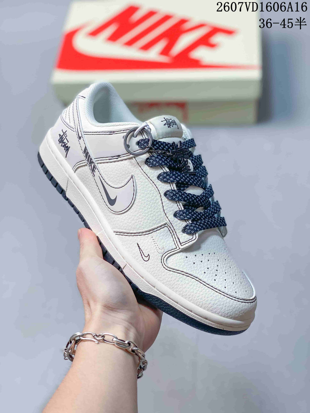 Close-up side view of a white low-top sneaker showing gray detailing, dark navy laces, perforated toe box, and decorative ring hardware.