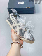 Angled side view of a silver sneaker with cream stripes and sheer bow laces.