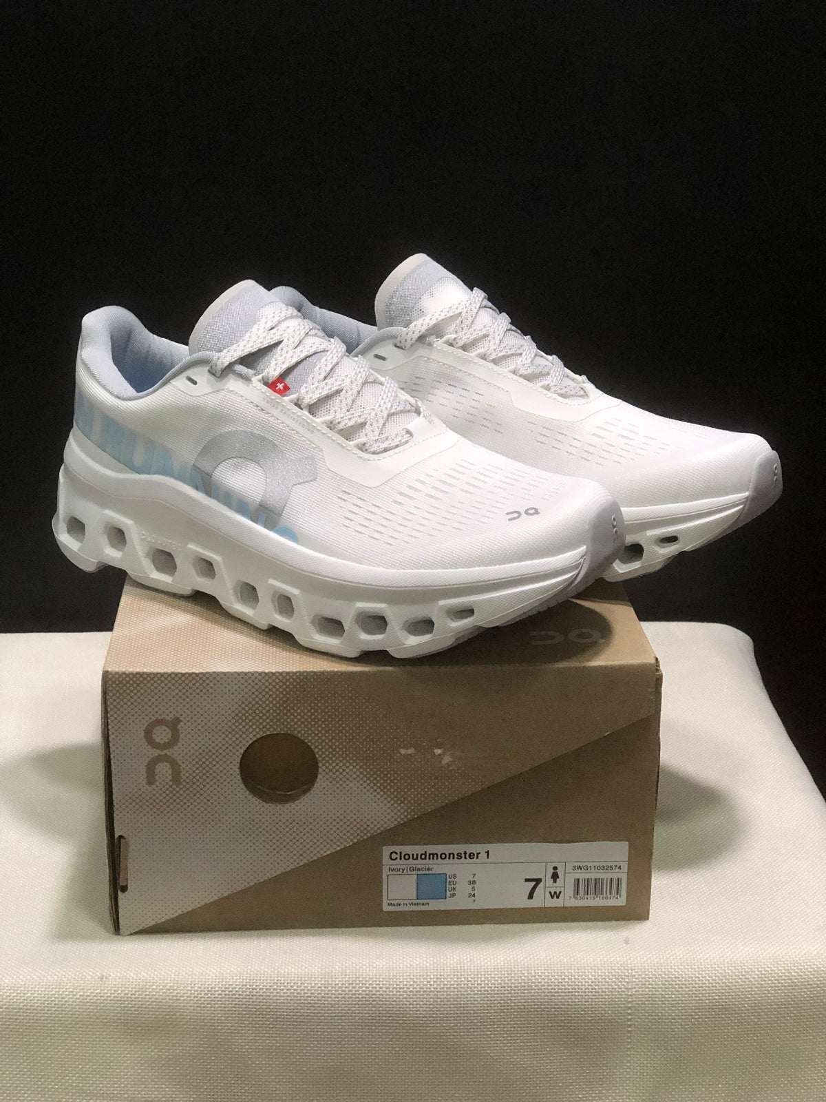 Pair of ivory and glacier On Cloudmonster 1 running shoes displayed on a branded shoe box; the box label shows EU 38 for the photographed sample.