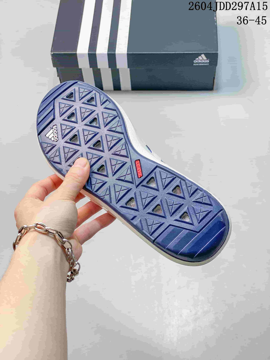 Close view of the blue patterned outsole with geometric tread openings and a small red outsole detail; an adidas shoebox is in the background.