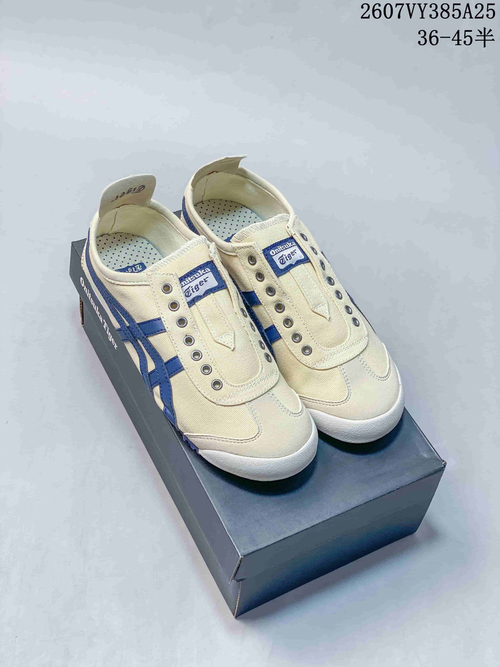 Pair of cream and blue Onitsuka Tiger slip-on sneakers displayed on a shoebox, with style code 2607VY385A25 and size range 36–45 shown above.