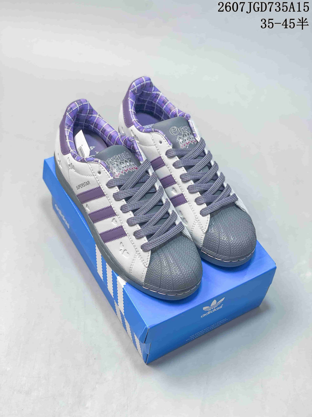 Pair of white Adidas Superstar sneakers with purple stripes, gray shell toes and a blue Adidas shoebox; image text shows style code 2607JGD735A15 and size range 35–45.