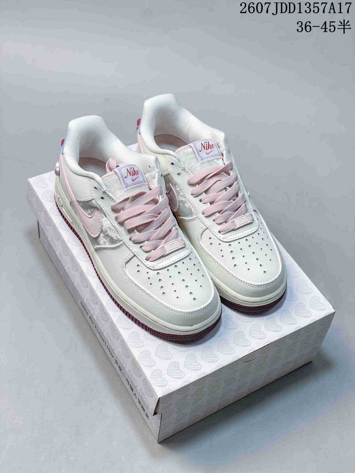 Pair of white Nike low-top sneakers with pink laces, blue details and mauve outsoles displayed on a heart-patterned box.