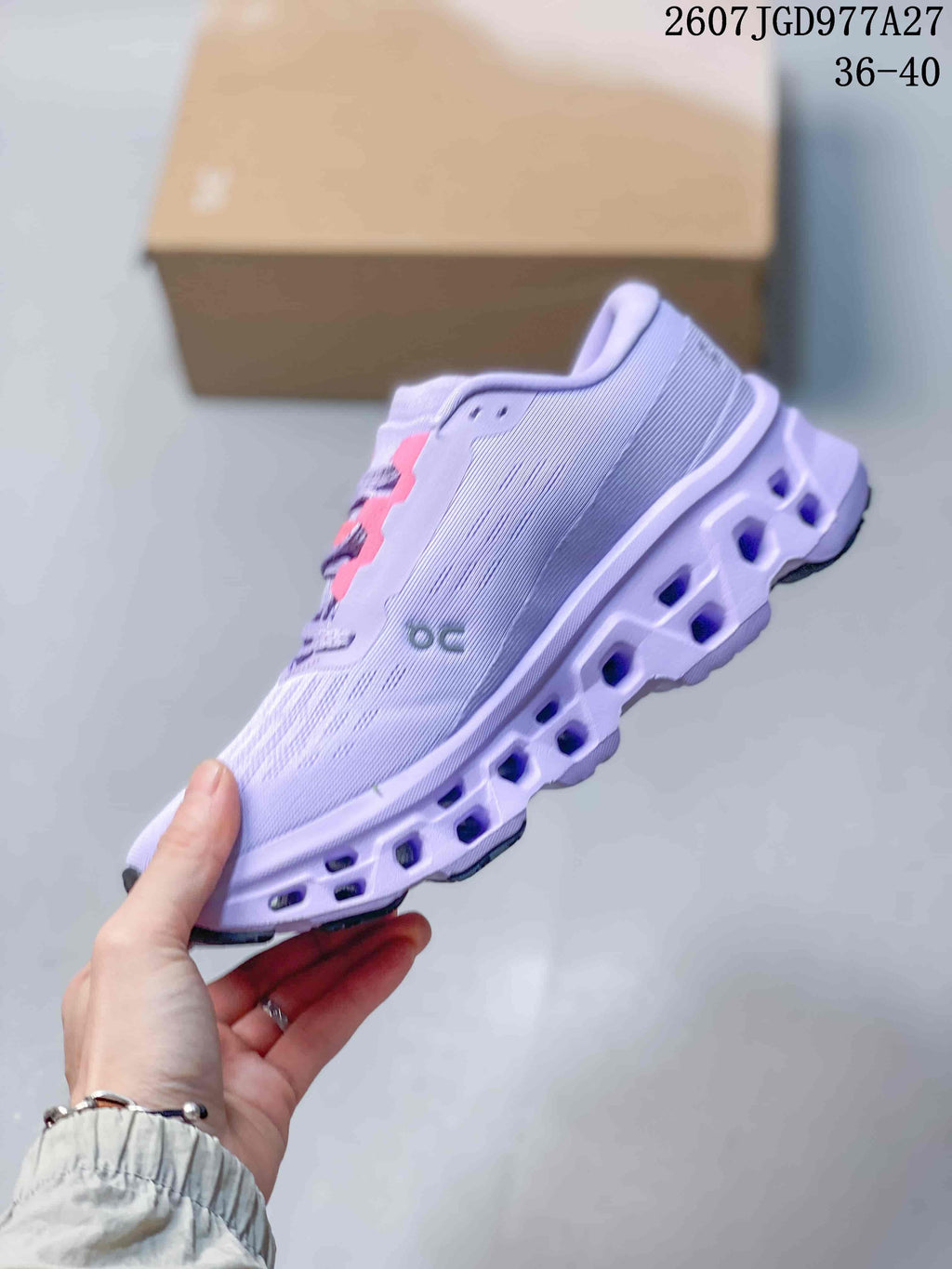 Side profile of a lavender On sneaker showing the textured upper, side logo, pink lace supports, and open segmented midsole; image text shows 2607JGD977A27 and 36–40.