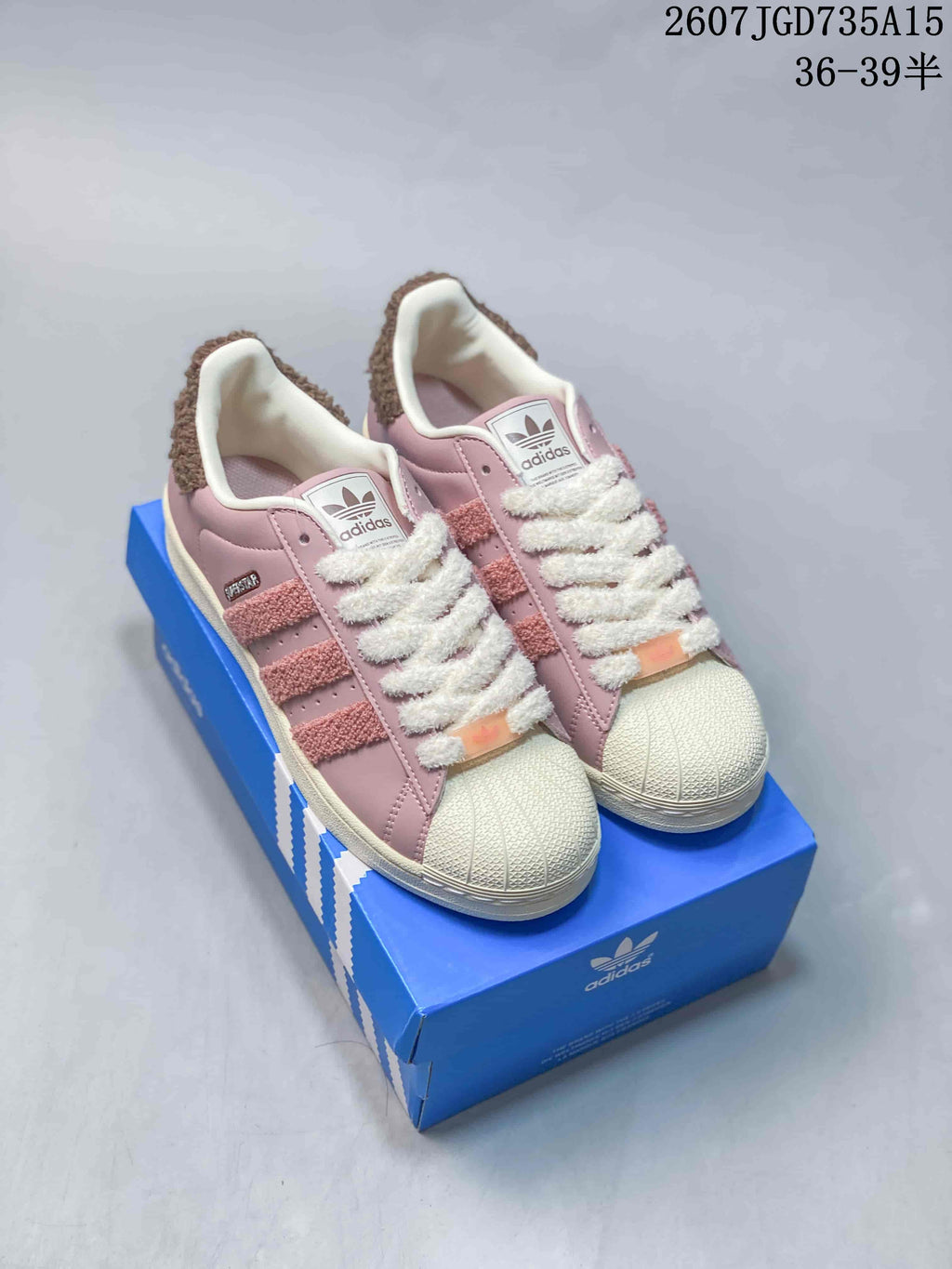 Pair of pink adidas Superstar-style sneakers displayed on a blue shoebox, showing the shell toes, textured laces and brown collar trim.