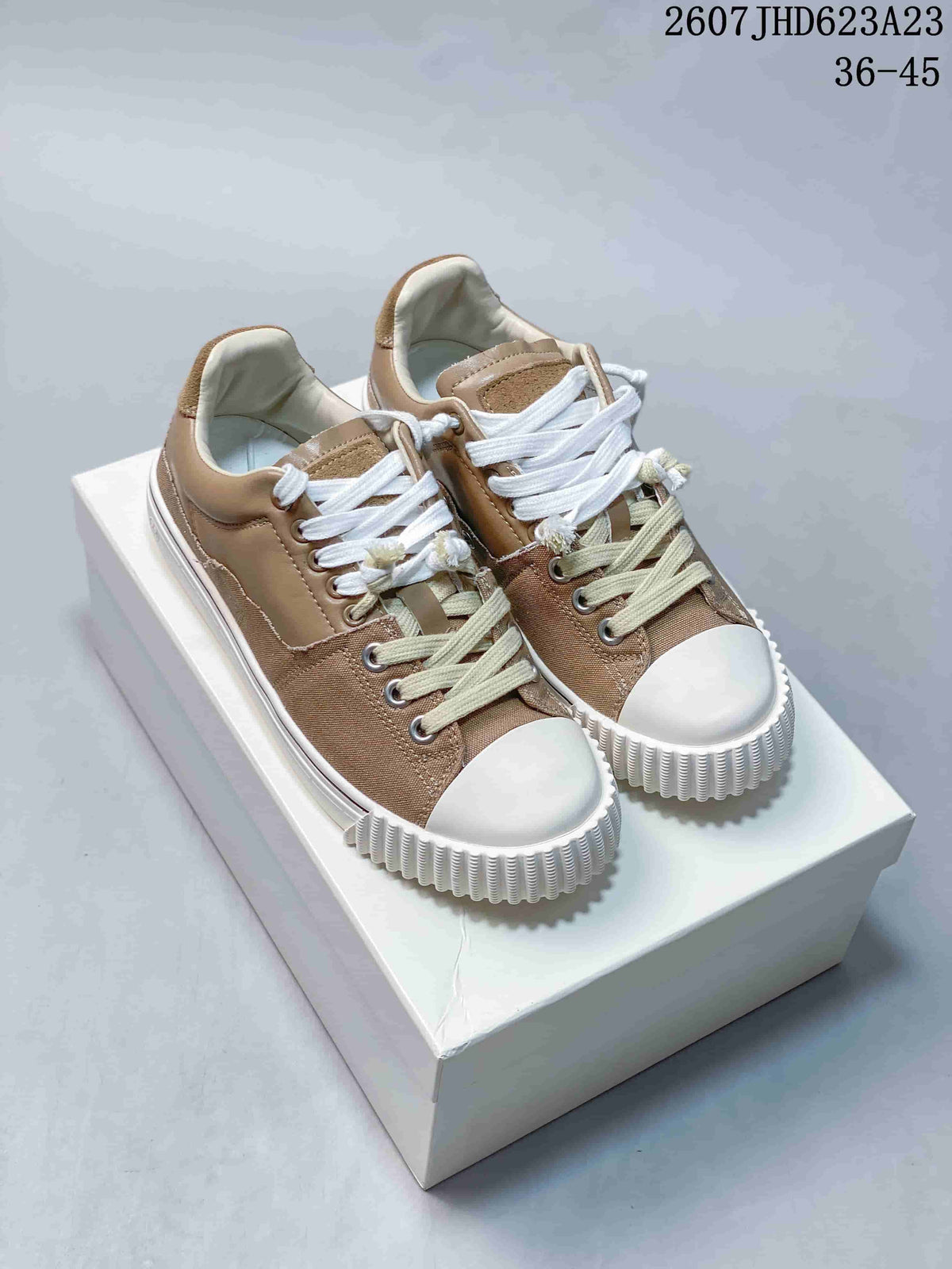 Pair of taupe low-top sneakers with white toe caps, contrasting laces, and thick white soles on a box.