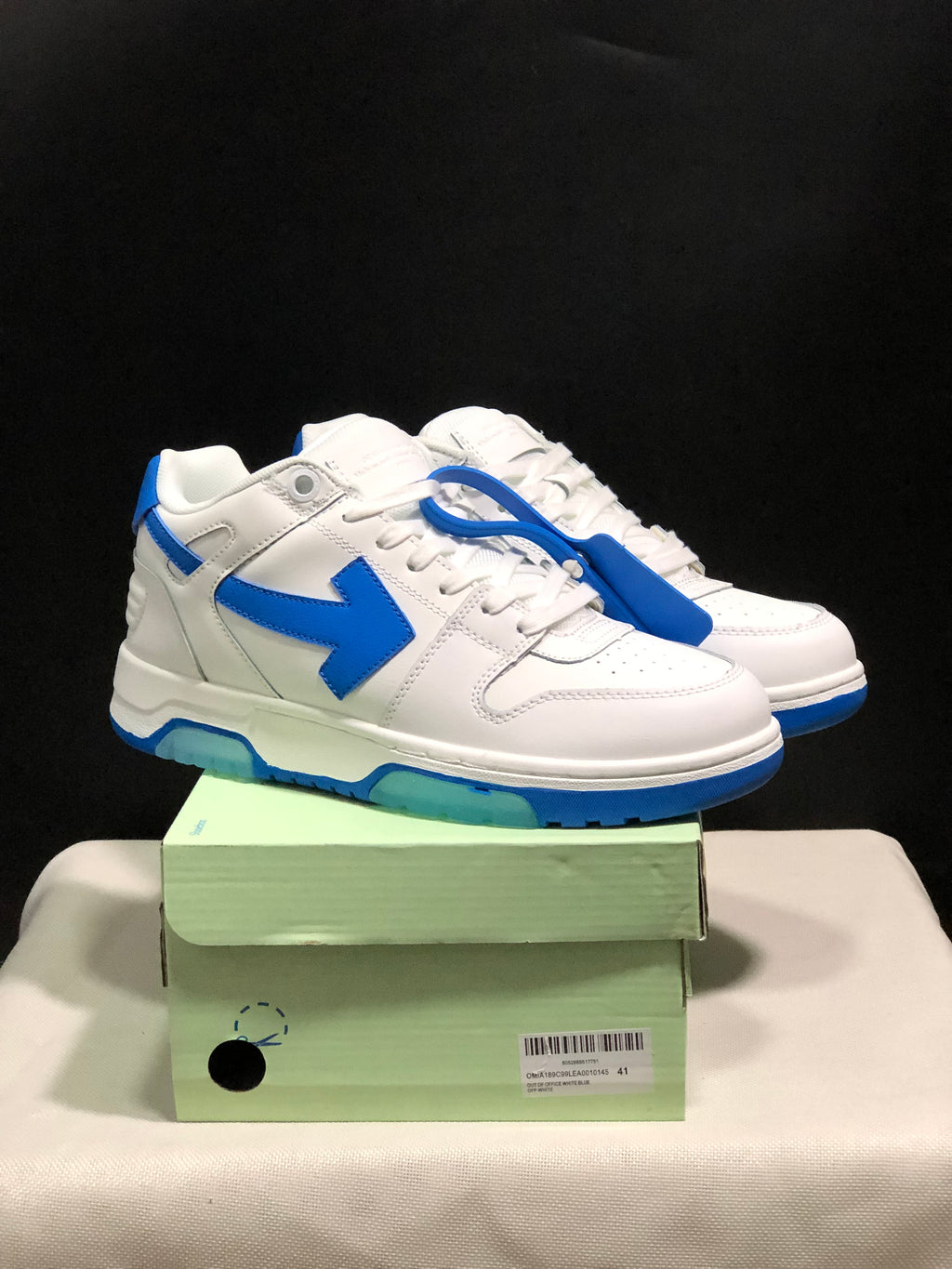 White and blue Off-White Out Of Office sneakers displayed on a shoebox, with a blue tag accessory visible.
