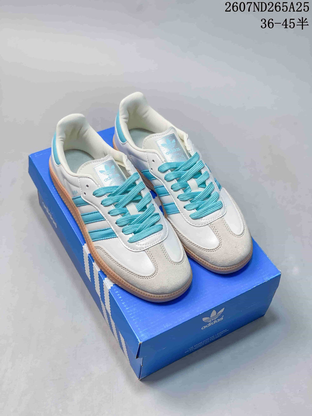 White adidas Samba sneakers with aqua accents and gum-toned outsoles displayed on a blue adidas shoebox.