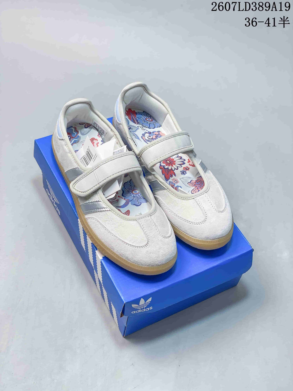 White adidas Originals Mary Jane sneakers with silver stripes, floral insoles, and gum-colored soles displayed on a blue shoebox.