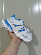 Angled close-up of the white and blue sneaker showing the adidas heel logo, layered side panels, blue collar, and side wording.
