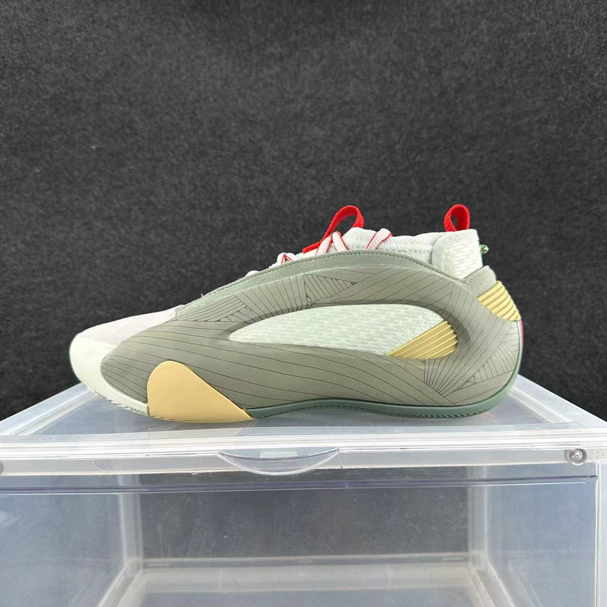 Side view of a low-top sneaker in pale gray-green and off-white, with red lace and pull-tab details and gold-toned accents.