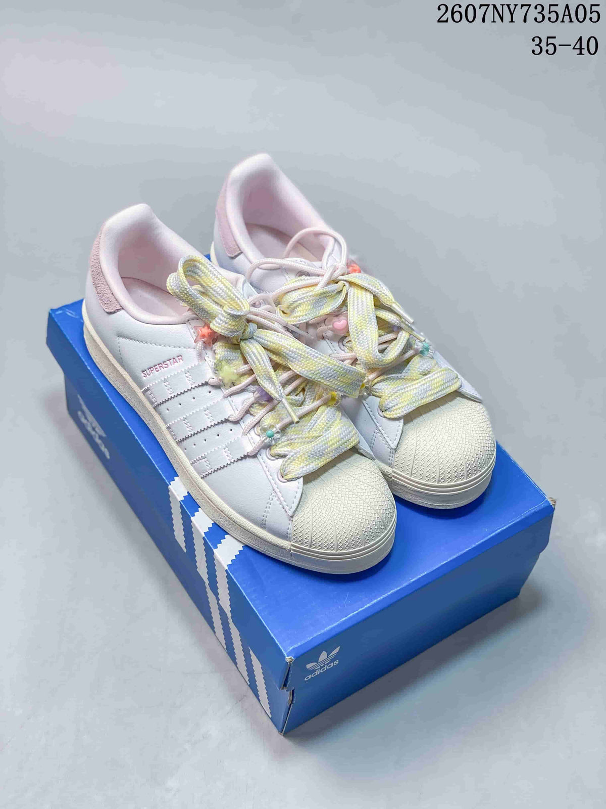 Pair of white Adidas Superstar sneakers with pastel decorative laces displayed on a blue Adidas shoe box.