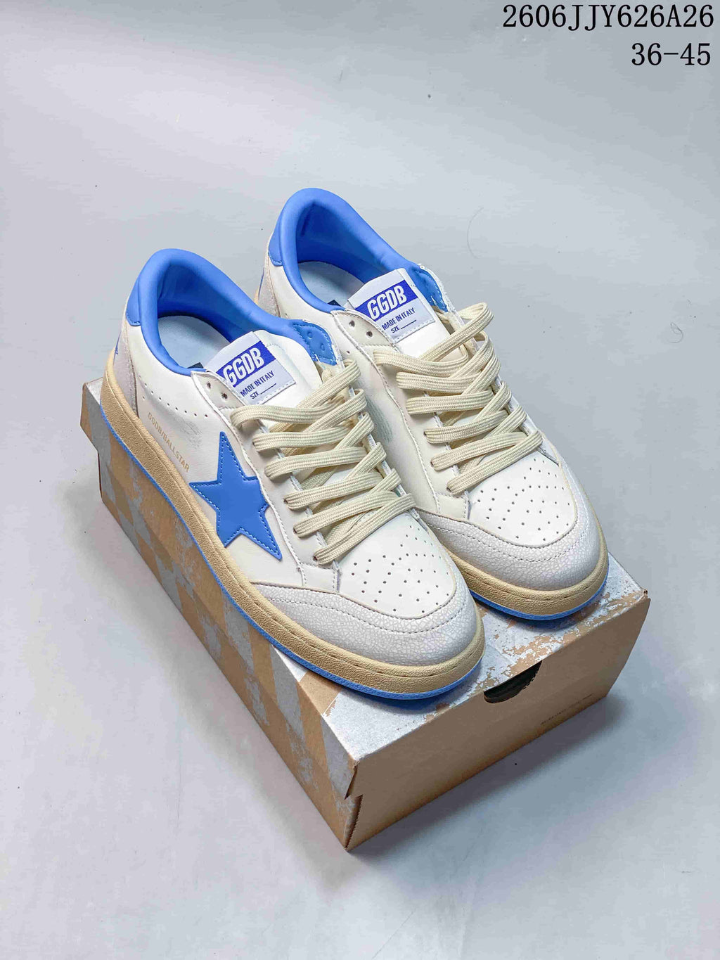 Pair of white and blue low-top sneakers with blue star details, cream laces, and a distressed beige-toned midsole displayed on a shoe box.