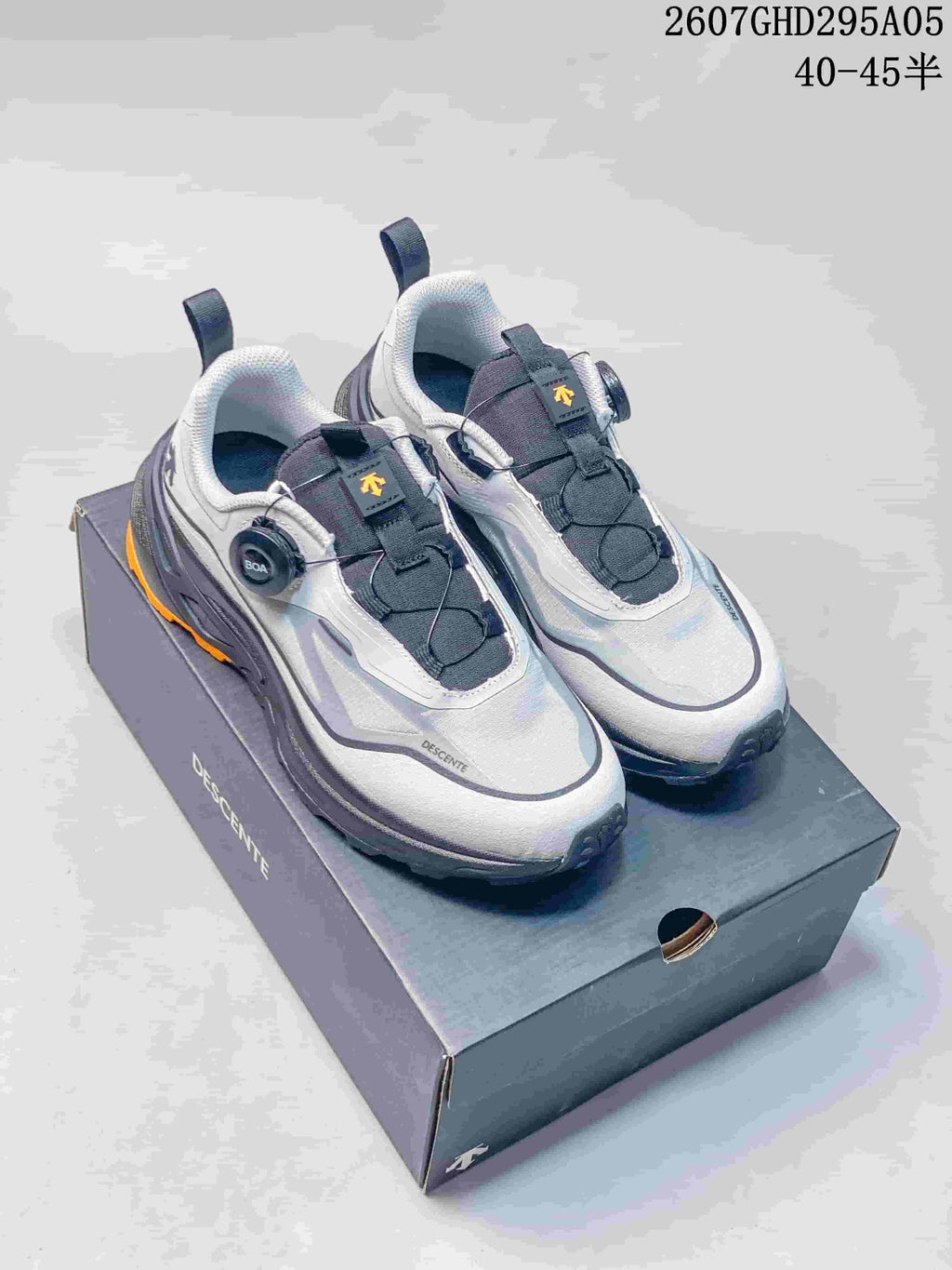Pair of white and gray Descente Fitstep Wire sneakers displayed on a branded shoebox, showing the BOA dials and orange outsole accents; style code 2607GHD295A05 and size range 40–45 are overlaid.