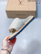 Angled side view of the ivory monogram-patterned slide sandal with tan trim and dark treaded outsole.