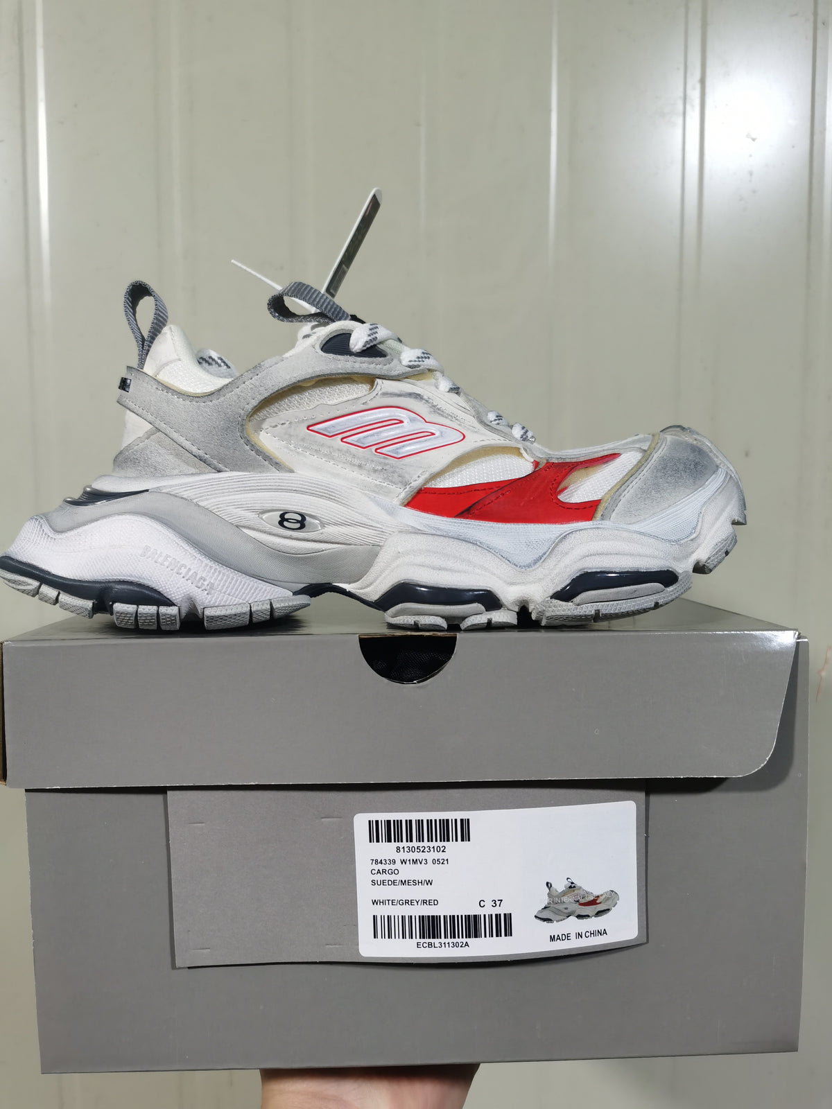 Side view of the white, grey and red sneaker on its box, with a visible label identifying style 784339 W1MV3 0521 and size C 37.
