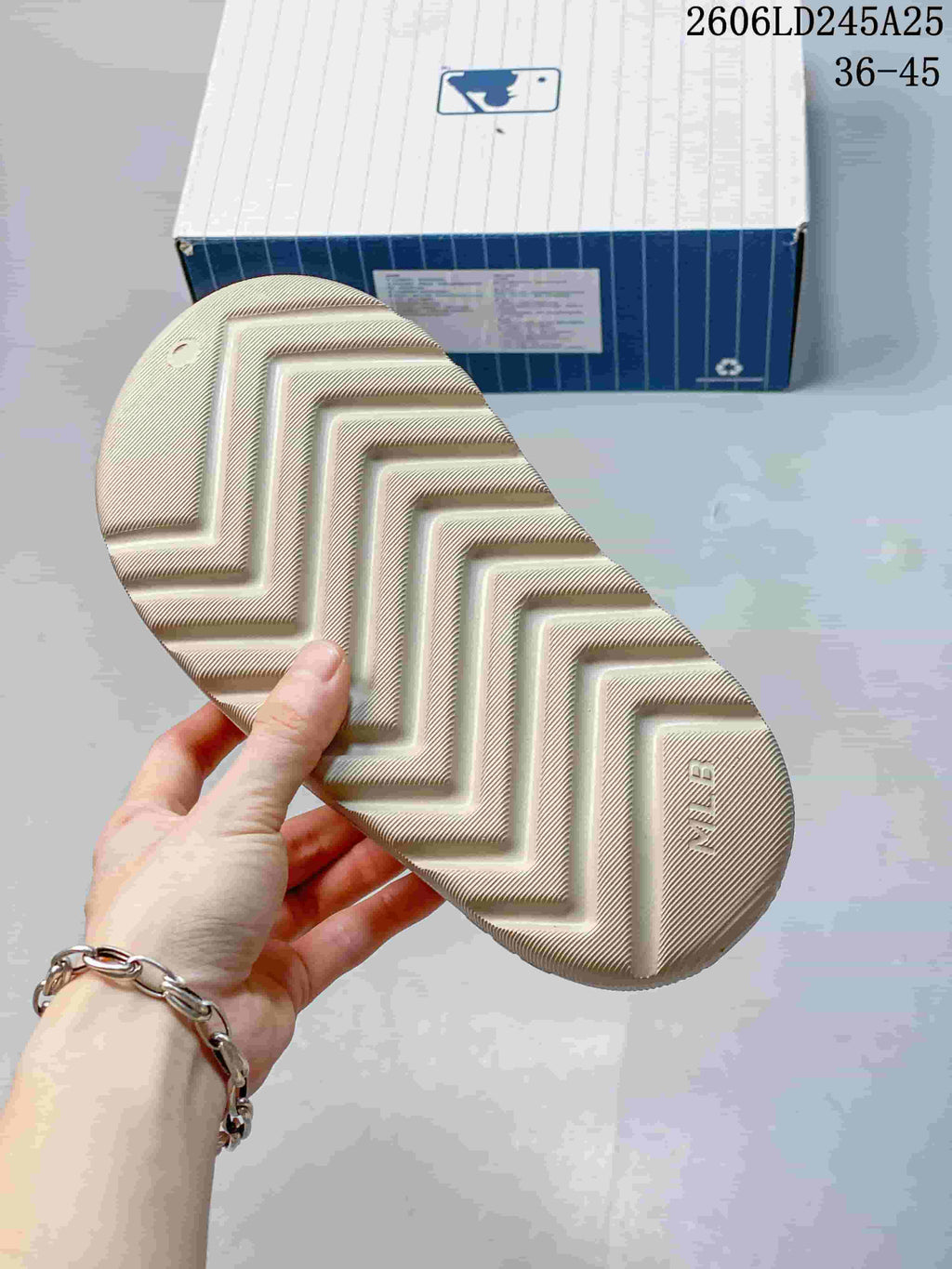 Underside of a beige slide sandal showing a textured zigzag outsole and MLB marking; the shoe box and size range 36-45 appear in the background.