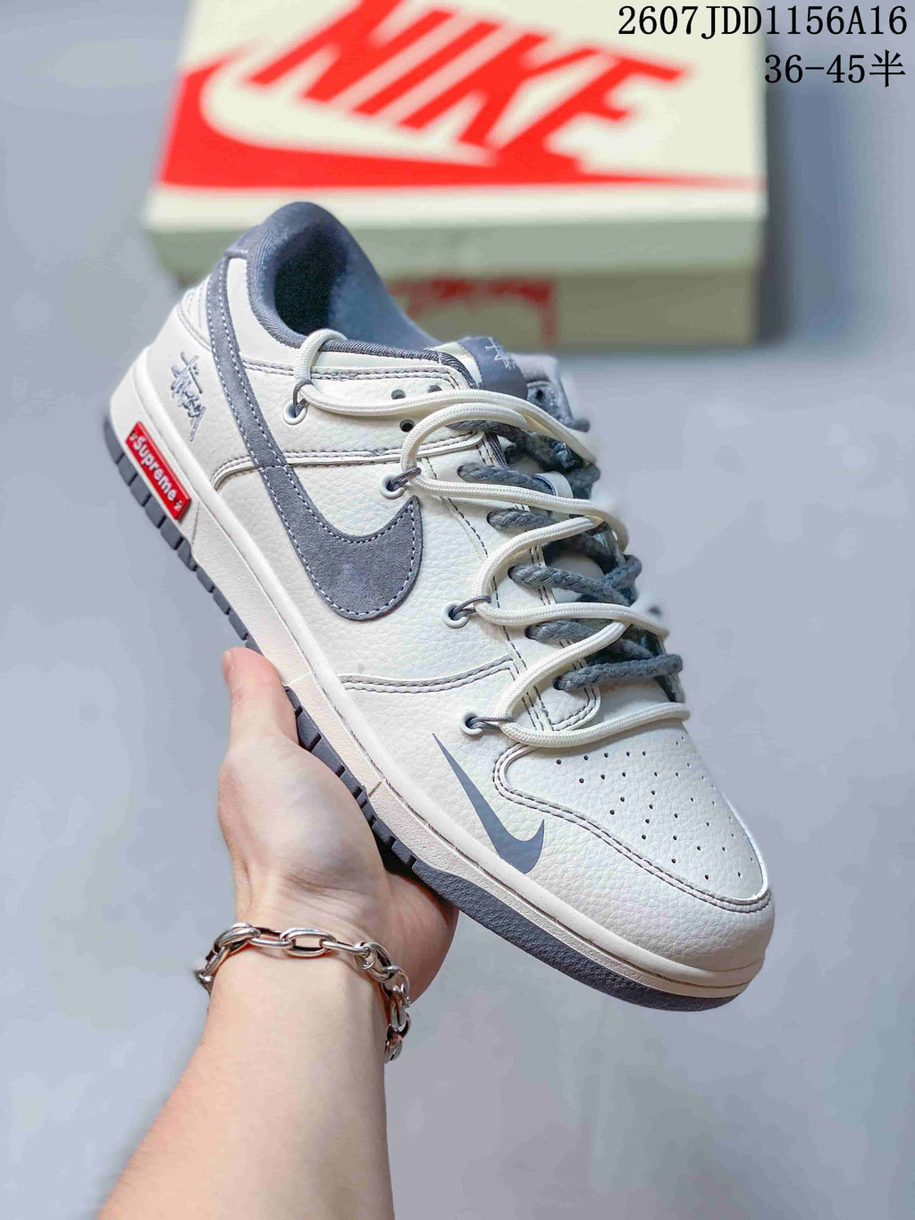Angled side view of an off-white and gray Nike SB Dunk Low sneaker with branded side details and a gray outsole; image text shows style code 2607JDD1156A16 and size range 36–45.5.