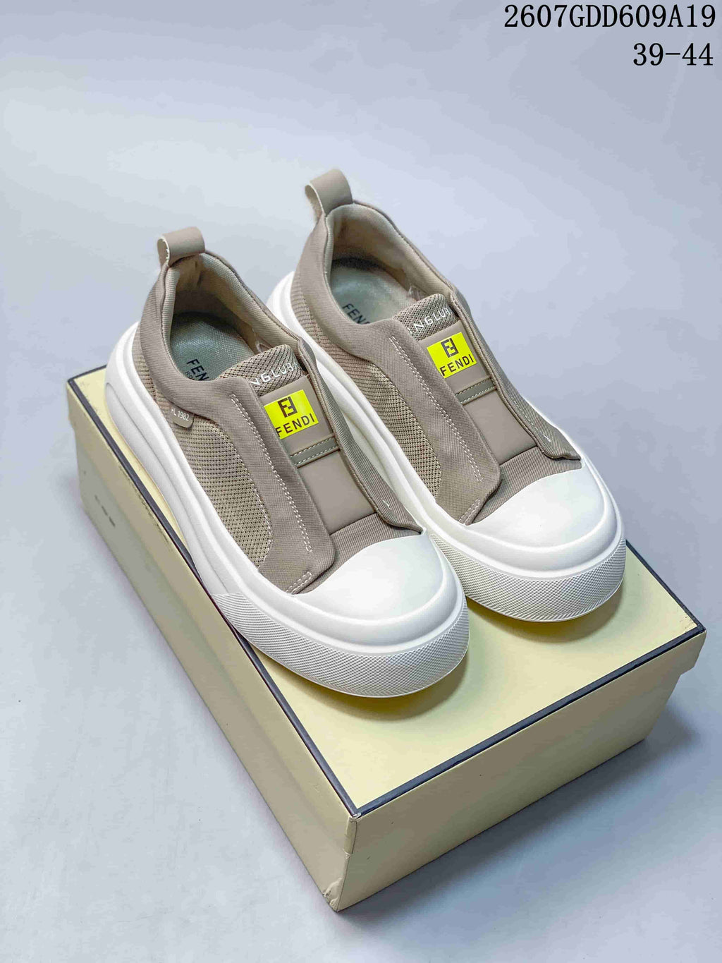 Taupe Fendi slip-on sneakers displayed on a shoebox, showing the front logo patches, pull tabs, and white soles; image text lists style code 2607GDD609A19 and sizes 39–44.