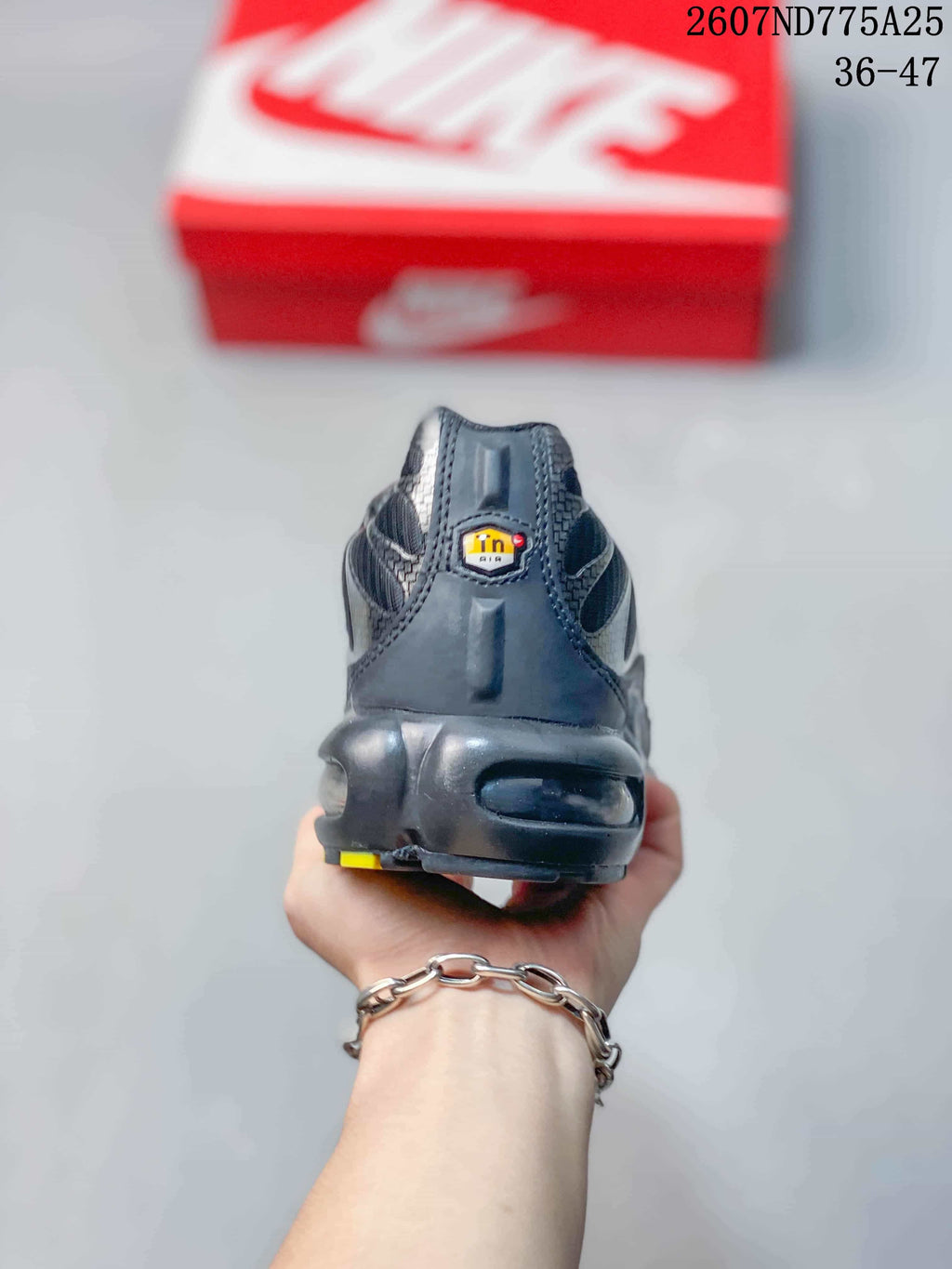 Rear view of a black Nike Air Max Plus TN sneaker showing the padded heel, outsole structure, and small yellow accent; overlay text shows code 2607ND775A25 and size range 36–47.