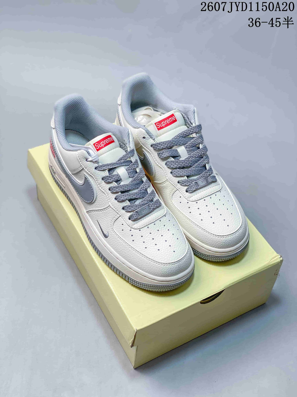 Pair of white and grey Nike Air Force 1 '07 low-top sneakers displayed on a pale yellow box; Supreme logo accents, product code 2607TYD1150A20, and the text 36-45 half sizes are visible.