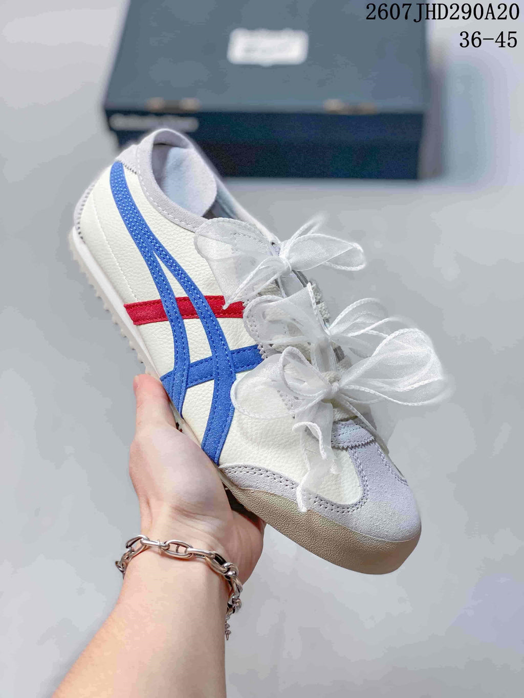 White sneaker with blue and red side stripes, gray toe detailing, and decorative ribbon laces.