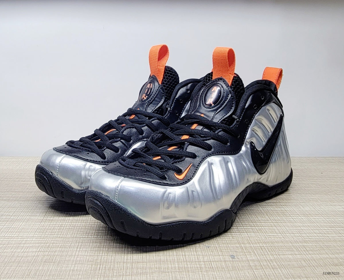 Pair of silver, black, and orange Nike Air Foamposite Pro sneakers viewed from the front and side