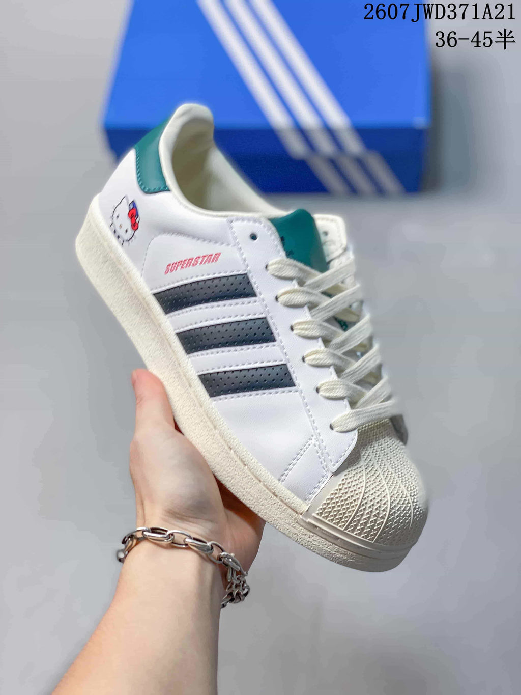Close-up of a white Adidas Superstar sneaker with dark blue striped side detailing, teal accents, a textured shell toe and visible Superstar branding; overlay states 2607TWD371A21 and sizes 36–45 with half sizes.