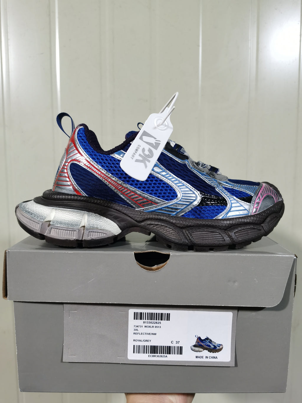 Royal blue, silver, black, grey, and pink low-top sneaker displayed on its box; the box label shows “REFLECTIVE/NW,” “ROYAL/GREY,” and size “C 37.”