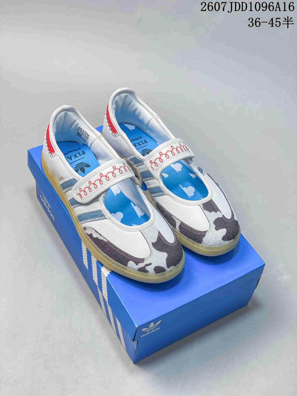 Pair of white adidas Samba Jane W sneakers with patterned toe panels, blue stripes, red accents, and a gum-colored outsole displayed on a blue shoebox.