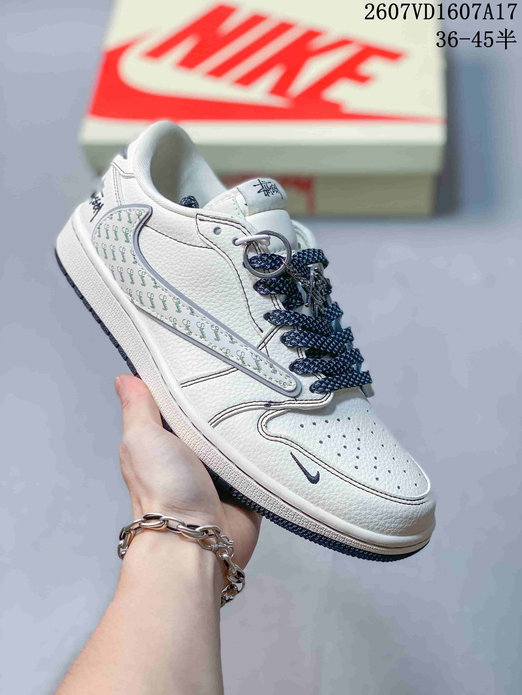 White low-top sneaker with repeating script-style graphics on the side overlay, dark laces and a small dark swoosh-style mark.