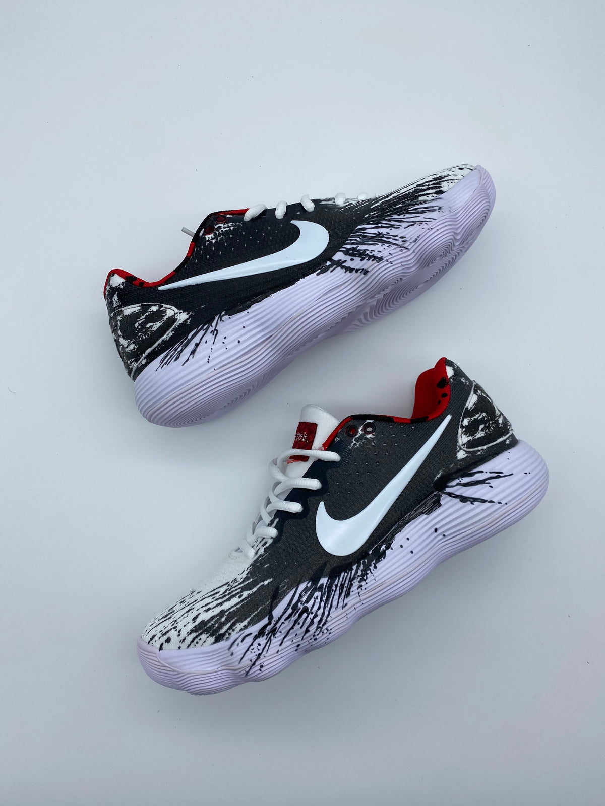 Pair of black Nike basketball shoes with white Swoosh logos, white laces, red collar accents, brushstroke graphics, and light-colored ridged outsoles.