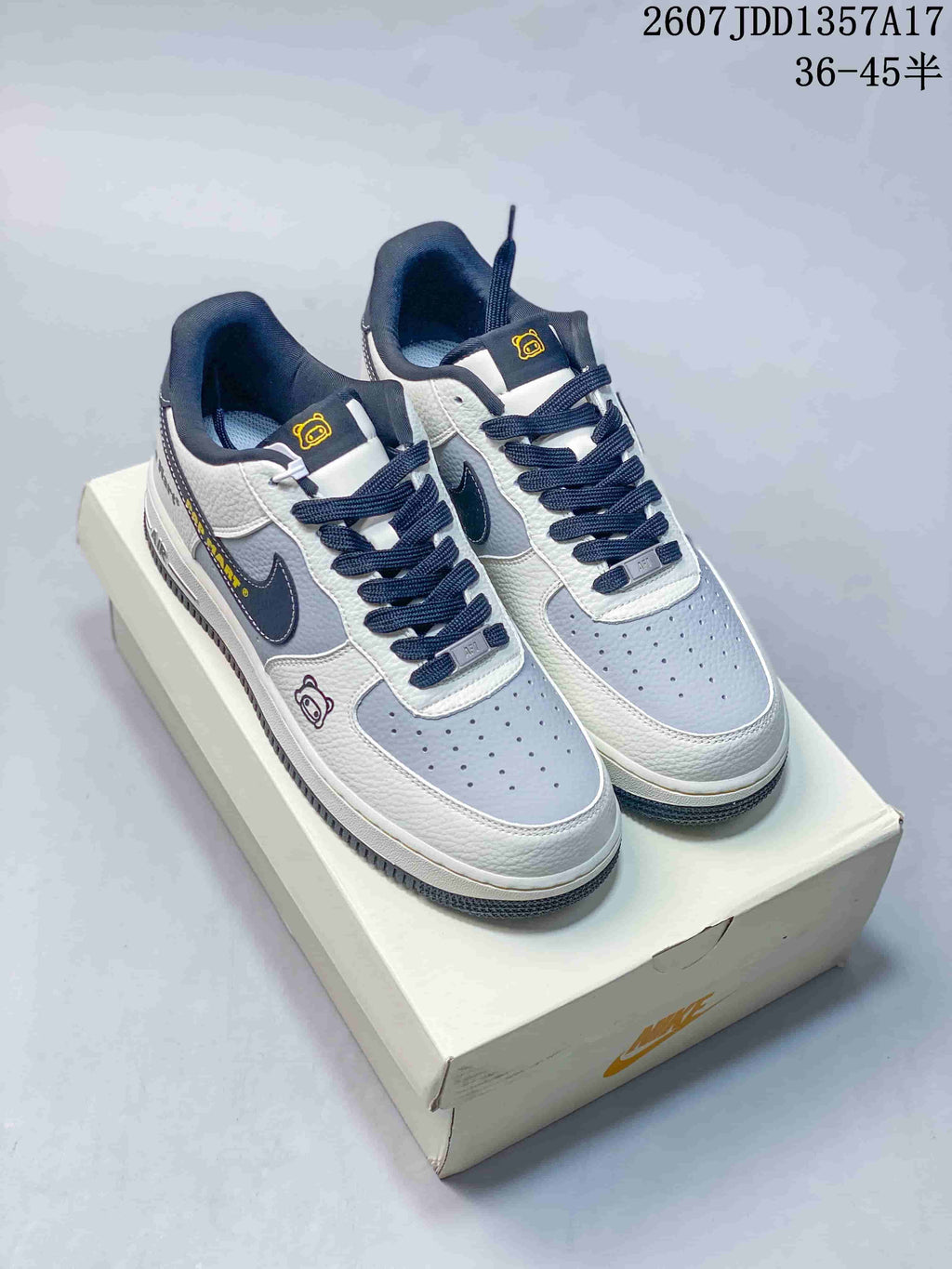 Pair of white, navy, and gray low-top sneakers displayed on a shoebox, showing dark laces and yellow tongue accents.