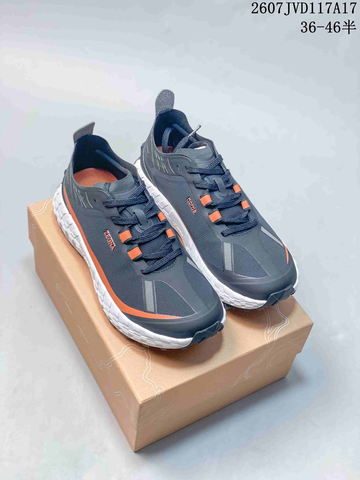 Pair of dark gray and black NORDA 001 G+ sneakers with orange accents and white midsoles displayed on a shoebox.