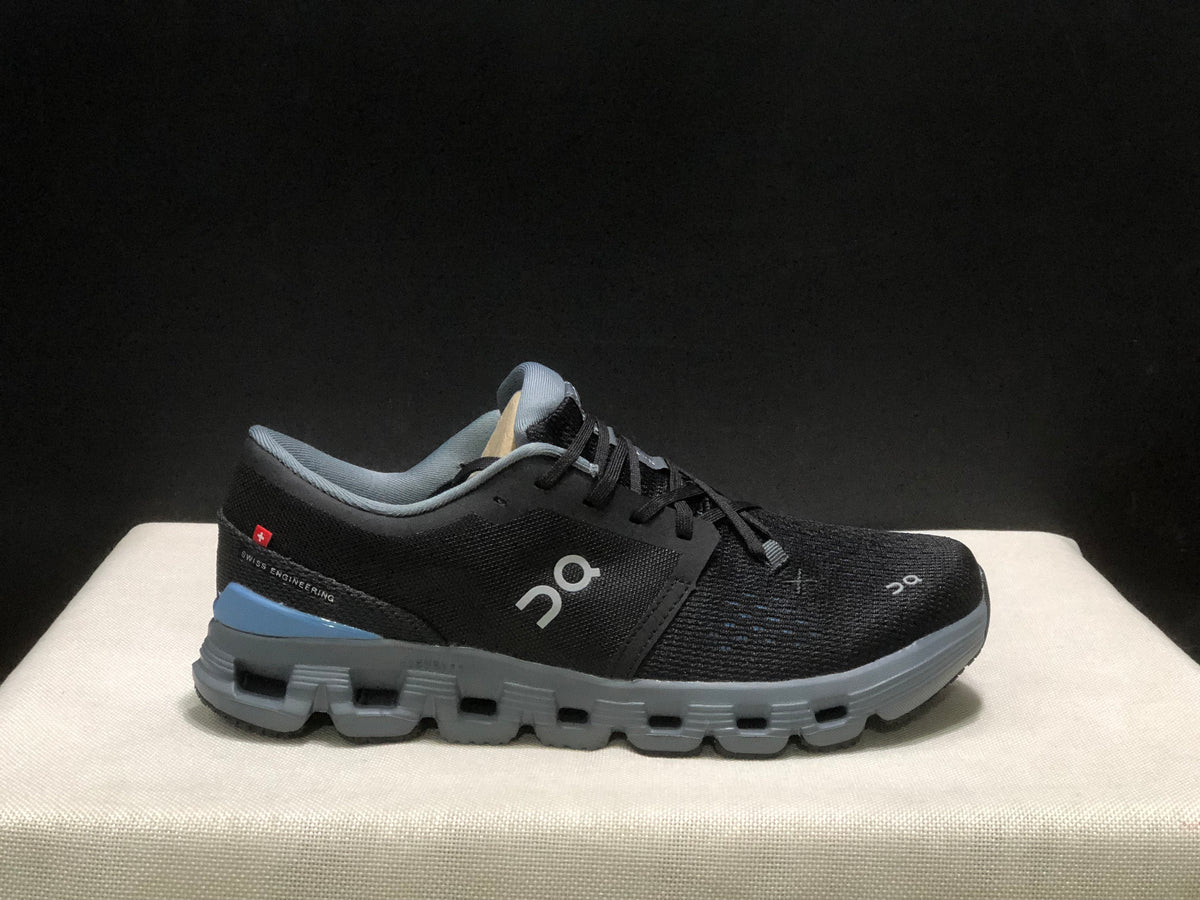 Side view of one black On Cloud X4 shoe showing the logo, gray sole, blue heel accent, and visible Swiss Engineering text.