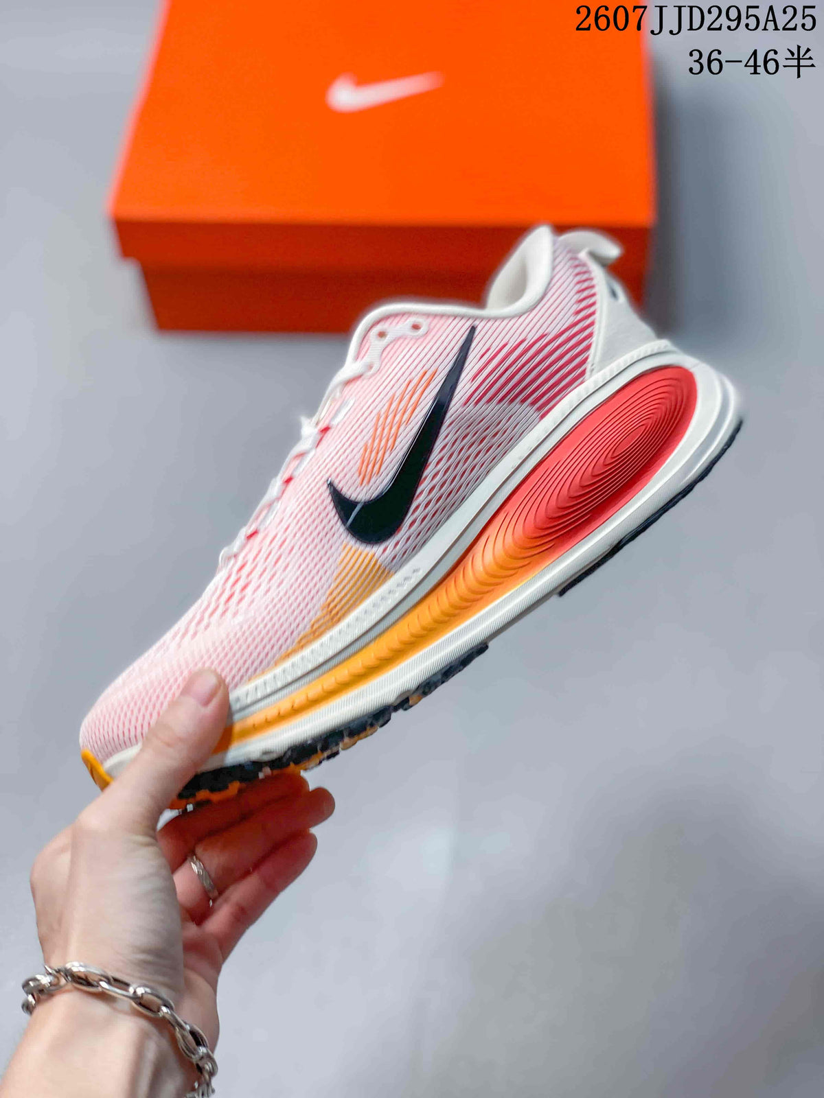 Side view of a pink and white Nike Vomero 18 sneaker showing the black Swoosh, orange accents, and raised midsole.