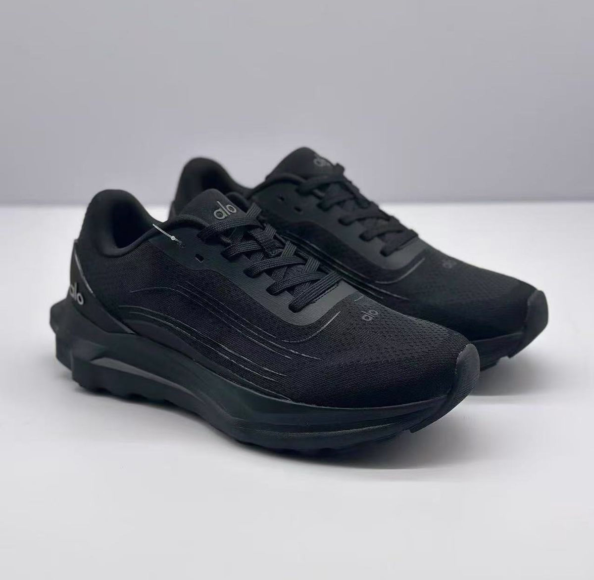 Front three-quarter view of a pair of all-black Alo sneakers with lace-up fronts and chunky soles.