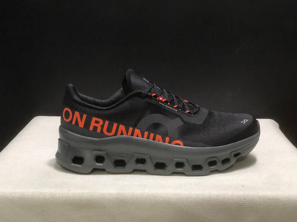 Side view highlighting the orange ON RUNNING graphic, black upper, and segmented outsole.