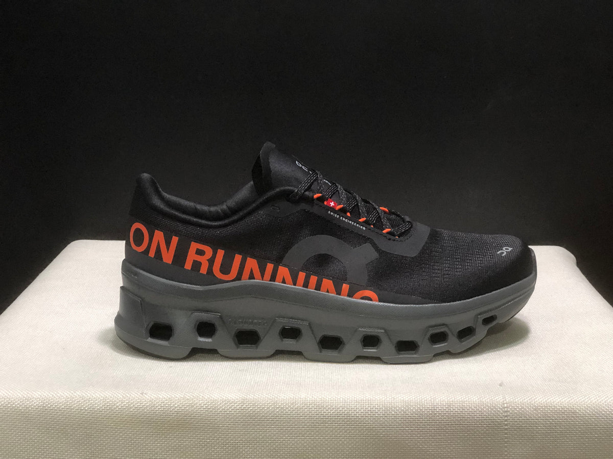 Side view highlighting the orange ON RUNNING graphic, black upper, and segmented outsole.
