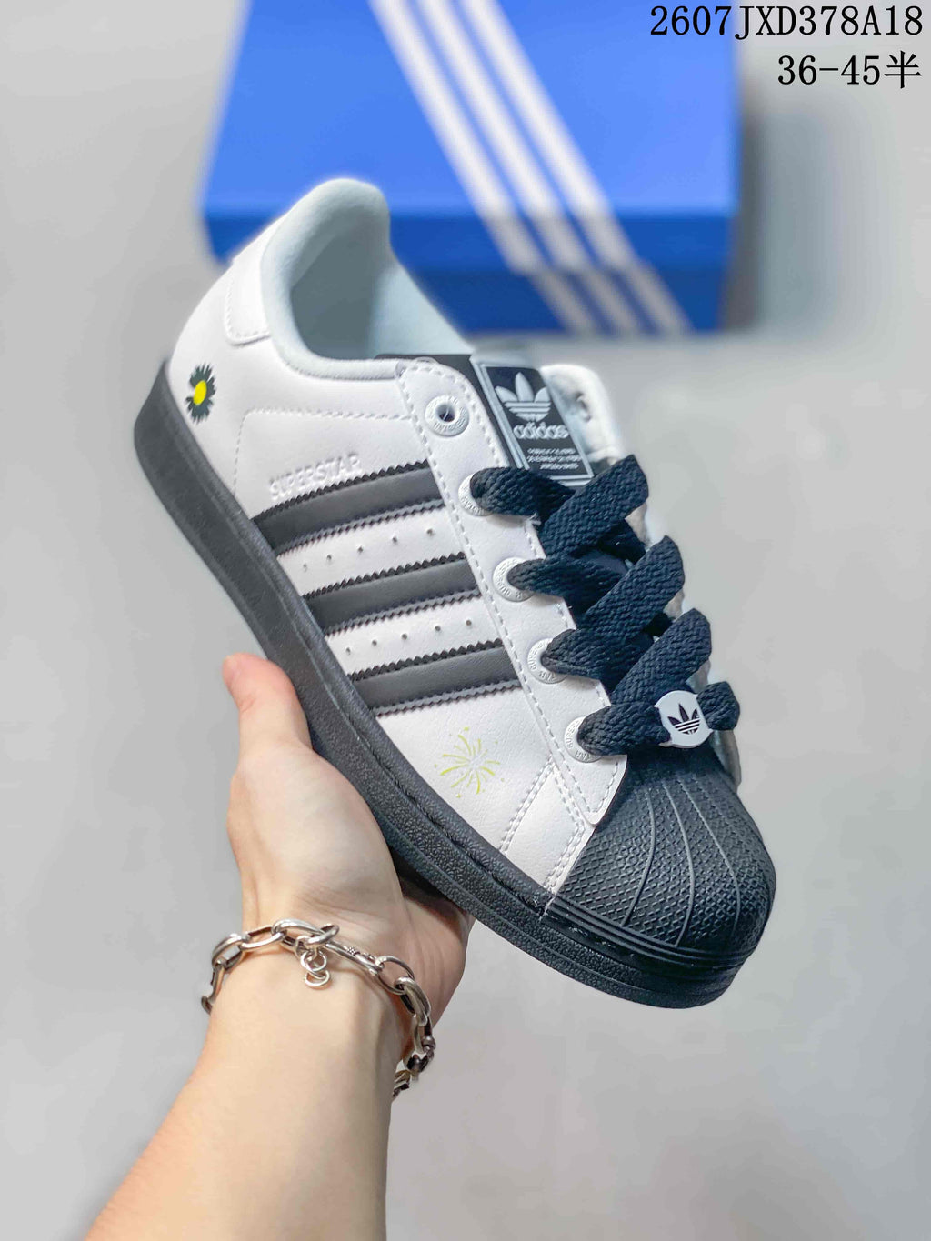 Angled side view of a white and black Superstar sneaker showing the side stripes, Superstar lettering, shell toe, and small daisy-style graphic.