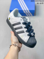 Angled side view of a white and black Superstar sneaker showing the side stripes, Superstar lettering, shell toe, and small daisy-style graphic.