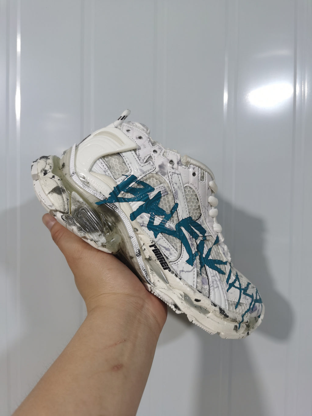 White and blue Balenciaga Runner sneaker held in hand, showing layered panels, mesh sections, and sculpted sole details.