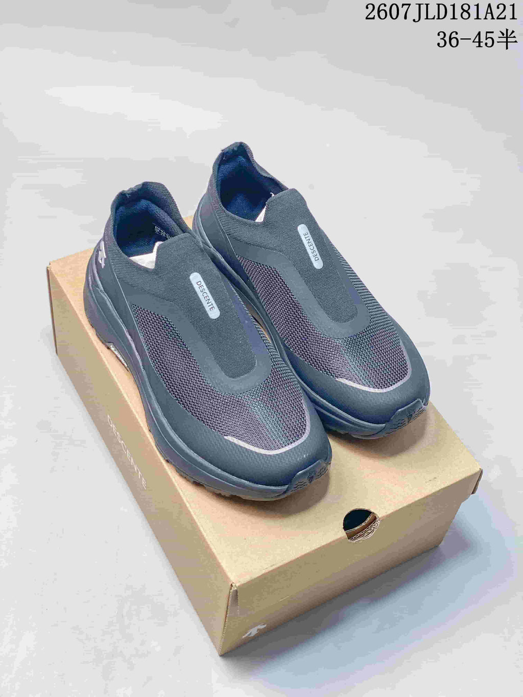 Pair of black DESCENTE PRIMER slip-on sneakers displayed on a shoe box, with the style code and size range visible above.