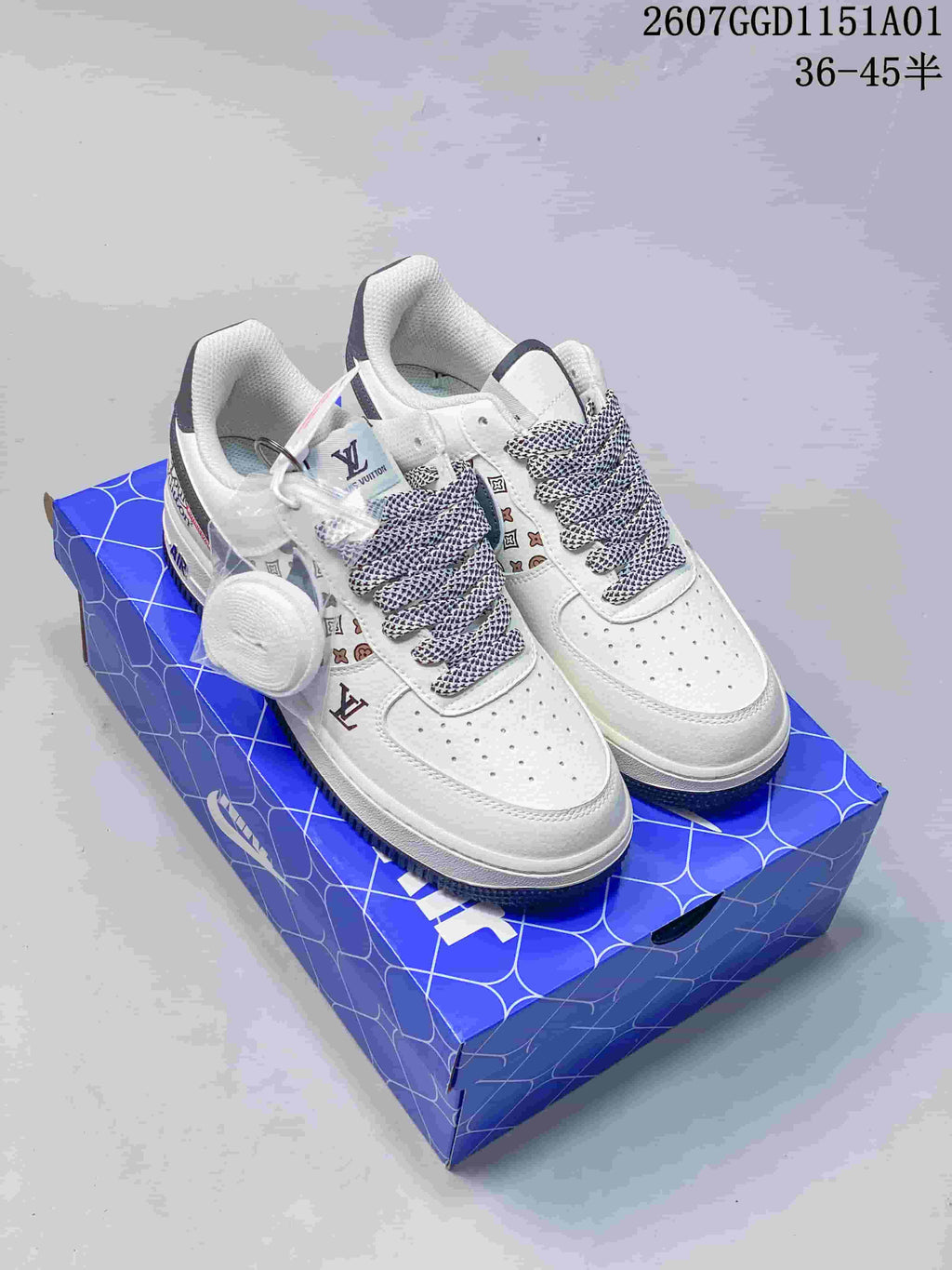 White low-top sneakers with patterned laces, monogram-style side print, and a round lace charm on a blue shoe box.