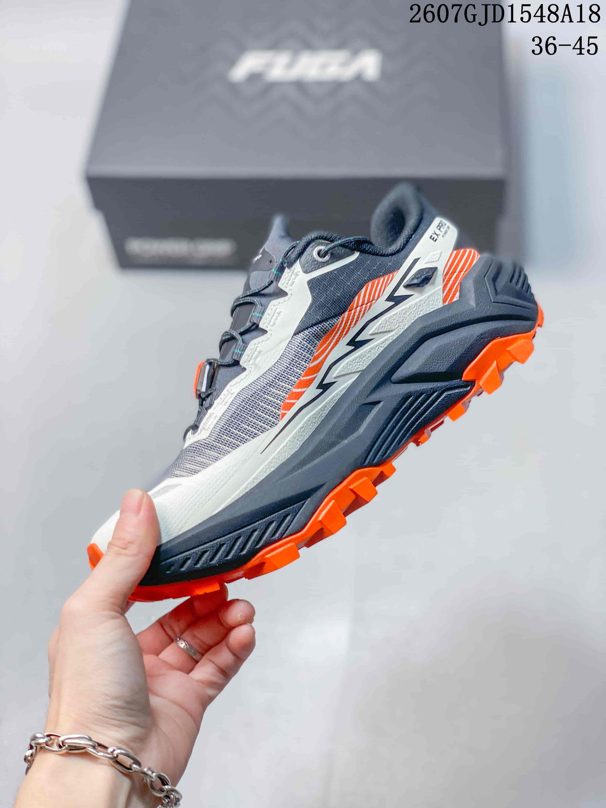 Side view highlighting the gray and white upper, layered midsole, orange outsole lugs, and FUGA heel branding.