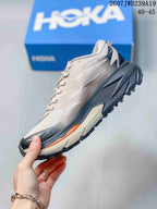 Side view of a low-top HOKA running shoe showing the light upper, dark sculpted midsole, orange accent, and lugged outsole.