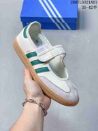 Side view of a white Samba Jane sneaker with green stripes, a hook-and-loop strap, and gum-colored outsole.