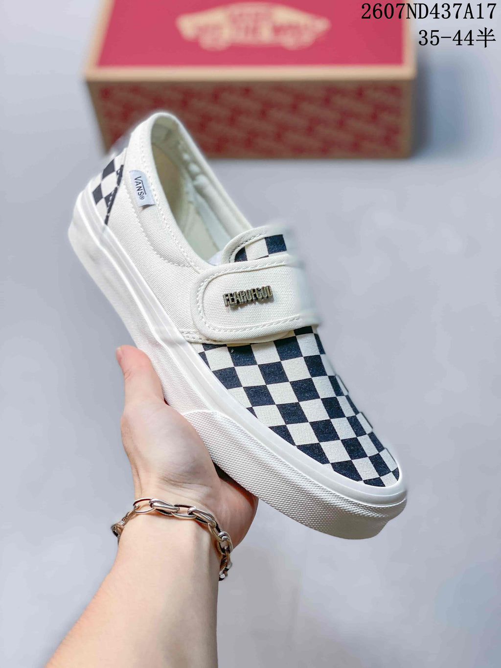 Side view of a black-and-white checkerboard Vans slip-on sneaker showing the strap, side branding, and textured white rubber sole.